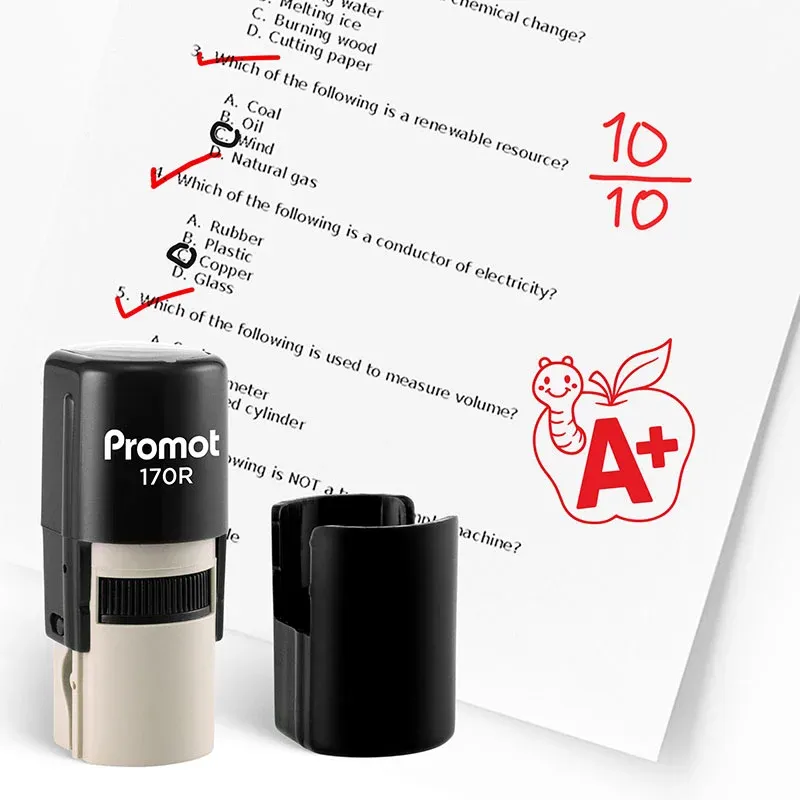 Black self inking teacher stamp and removable cap displayed beside a graded science worksheet marked with red check marks and a “10/10” score. A red apple stamp with a worm and “A+” design appears on the page, showing how the classroom stamp can reward ex