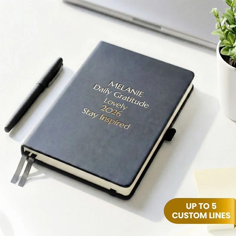 Personalized leather journal with a black faux leather cover, custom gold foil text, ribbon bookmarks, and an elastic closure, displayed on a white desk beside a black pen and a small potted plant.