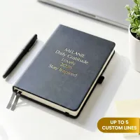 Personalized leather journal with a black faux leather cover, custom gold foil text, ribbon bookmarks, and an elastic closure, displayed on a white desk beside a black pen and a small potted plant.