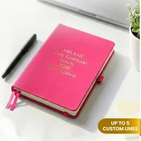 Pink journal with gold engraved text on a desk beside a pen, a personalized leather journal with up to 5 custom lines.