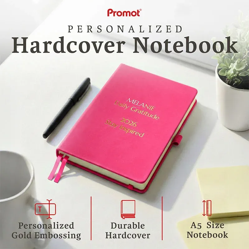 Pink hardcover notebook with gold embossed text on a desk, a personalized leather journal with custom engraving and ribbon bookmarks.