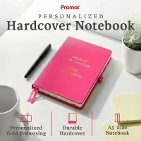 Pink hardcover notebook with gold embossed text on a desk, a personalized leather journal with custom engraving and ribbon bookmarks.