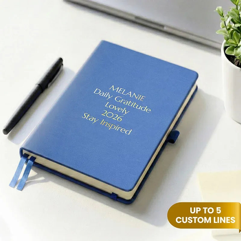 Blue personalized leather journal with custom gold embossed text, displayed on a white desk beside a pen and potted plant.