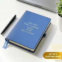 Blue personalized leather journal with custom gold embossed text, displayed on a white desk beside a pen and potted plant.