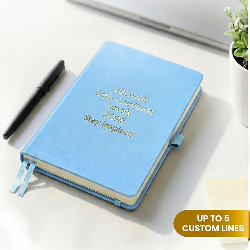 Sky blue notebook with custom gold engraving on a desk beside a pen, a personalized leather journal with two ribbon bookmarks.