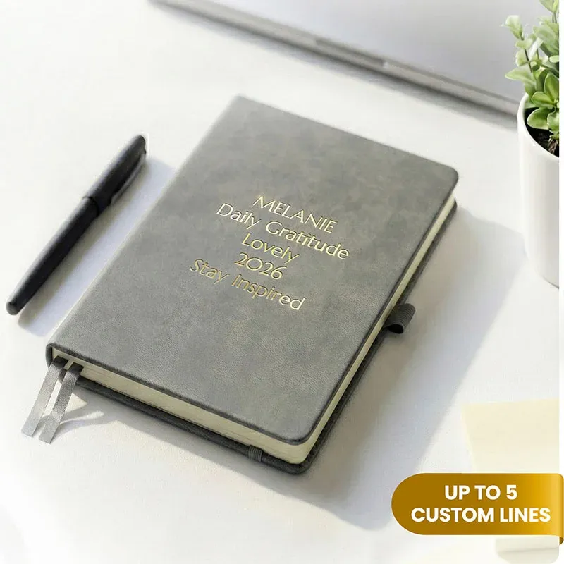 Gray journal with gold engraved text on a desk beside a pen, a personalized leather journal with up to 5 custom lines.