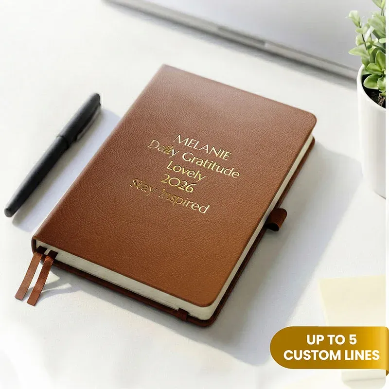 Brown personalized leather journal with custom gold embossed text, displayed on a white desk beside a pen and potted plant.