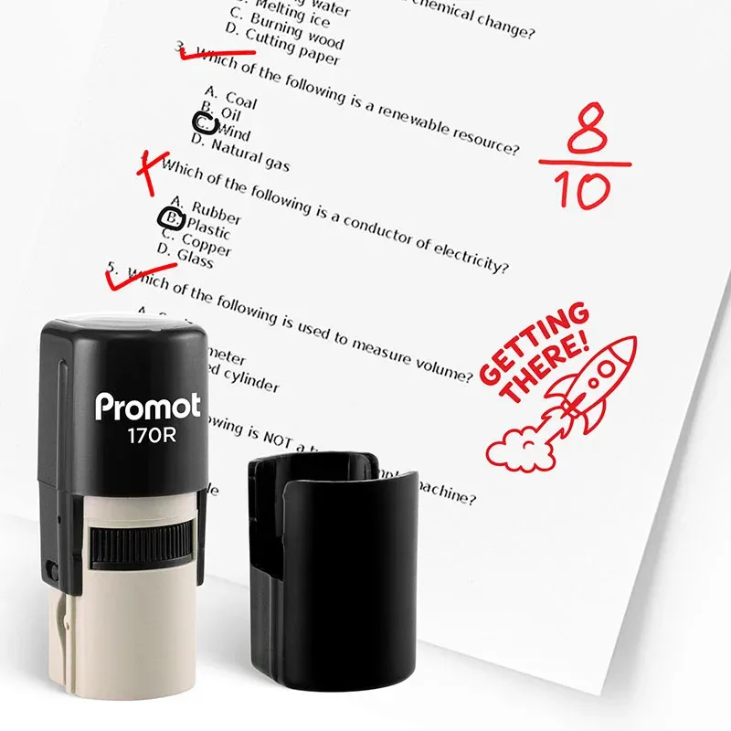 Black self inking stamp and removable cap displayed beside a graded science worksheet marked with red check marks, an “8/10” score, and a rocket stamp that says “GETTING THERE!” The worksheet includes multiple choice science questions, and this ink stamp