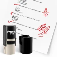 Black self inking stamp and removable cap displayed beside a graded science worksheet marked with red check marks, an “8/10” score, and a rocket stamp that says “GETTING THERE!” The worksheet includes multiple choice science questions, and this ink stamp