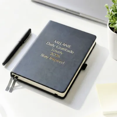Personalized leather journal with a black faux leather cover, custom gold foil text, ribbon bookmarks, and an elastic closure, displayed on a white desk beside a black pen and a small potted plant.