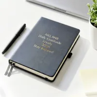 Personalized leather journal with a black faux leather cover, custom gold foil text, ribbon bookmarks, and an elastic closure, displayed on a white desk beside a black pen and a small potted plant.
