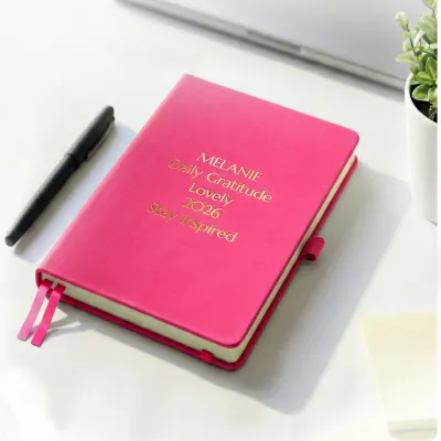 Pink journal with gold engraved text on a desk beside a pen, a personalized leather journal with up to 5 custom lines.