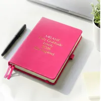 Pink journal with gold engraved text on a desk beside a pen, a personalized leather journal with up to 5 custom lines.