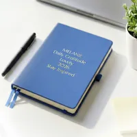 Blue personalized leather journal with custom gold embossed text, displayed on a white desk beside a pen and potted plant.