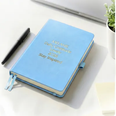 Sky blue notebook with custom gold engraving on a desk beside a pen, a personalized leather journal with two ribbon bookmarks.