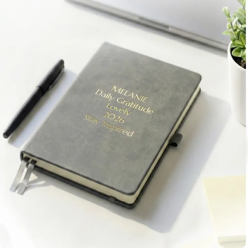 Gray journal with gold engraved text on a desk beside a pen, a personalized leather journal with up to 5 custom lines.