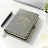 Gray journal with gold engraved text on a desk beside a pen, a personalized leather journal with up to 5 custom lines.