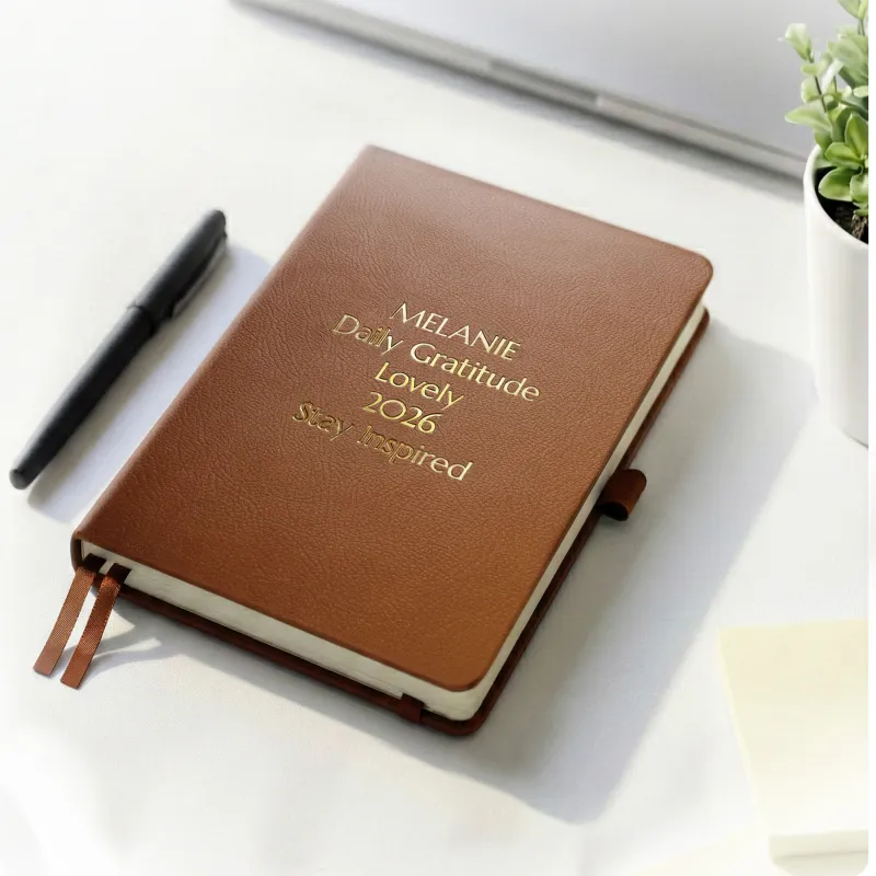 Brown personalized leather journal with custom gold embossed text, displayed on a white desk beside a pen and potted plant.