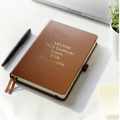Brown personalized leather journal with custom gold embossed text, displayed on a white desk beside a pen and potted plant.